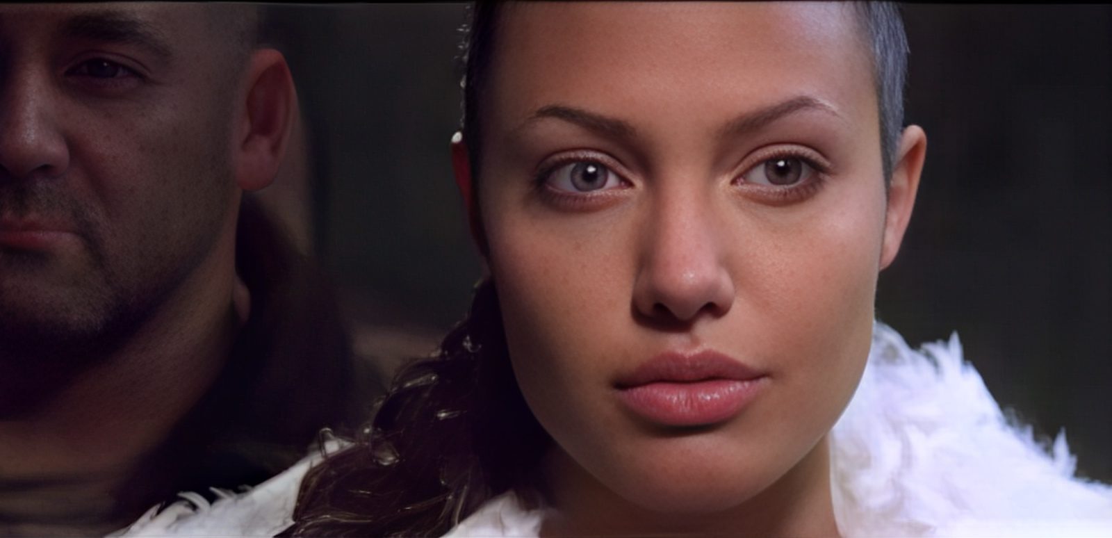 A woman with light eyes and a white fur collar looks forward, while a man stands behind her in the shadows, partially obscured.