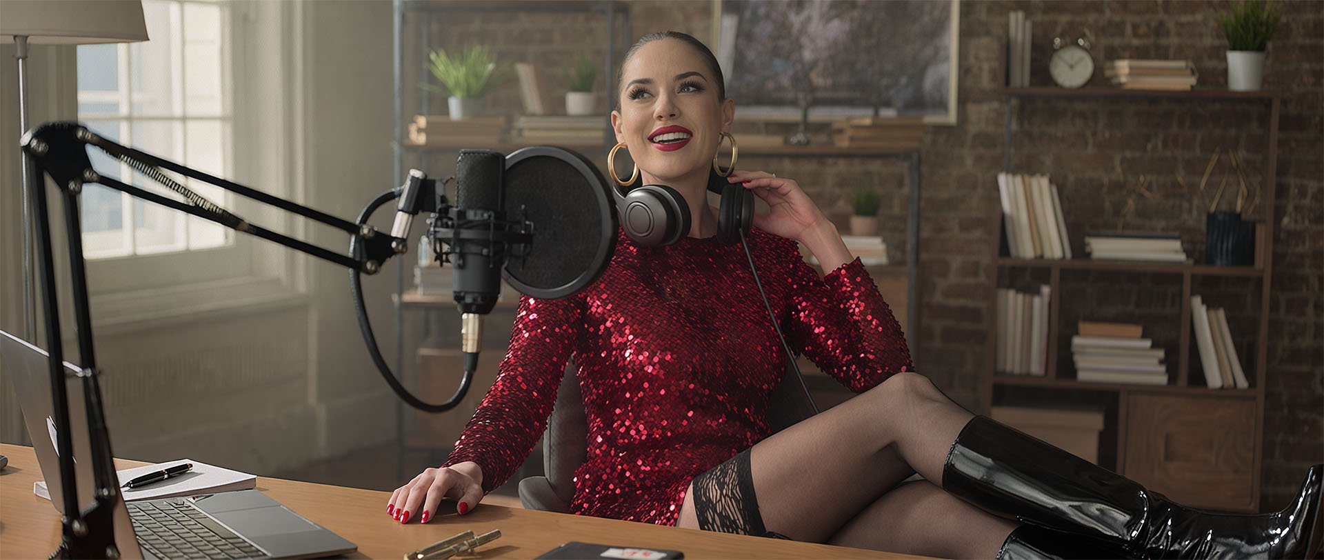 A woman in a red sequin dress and black boots sits at a desk with a microphone and headphones, smiling in a modern recording studio with bookshelves in the background.