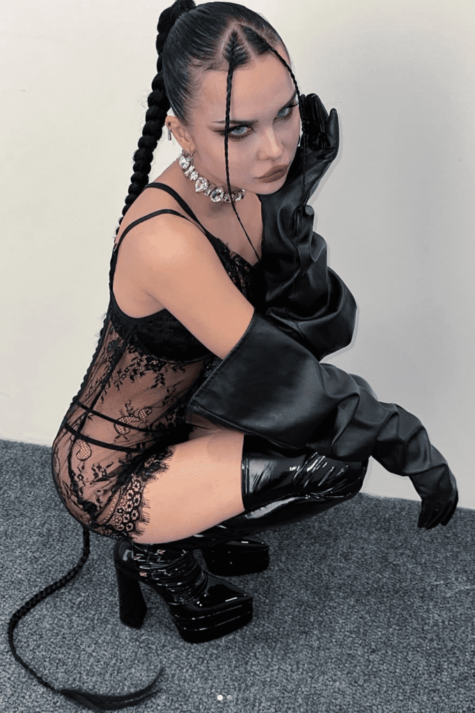 Person wearing black lace bodysuit, long gloves, high platform boots, and large necklace squats on a carpeted floor against a plain wall, with hair styled in long braids.