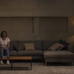 A woman in casual clothes sits on a couch while another woman in a shiny dress and heels stands facing her in a modern living room.
