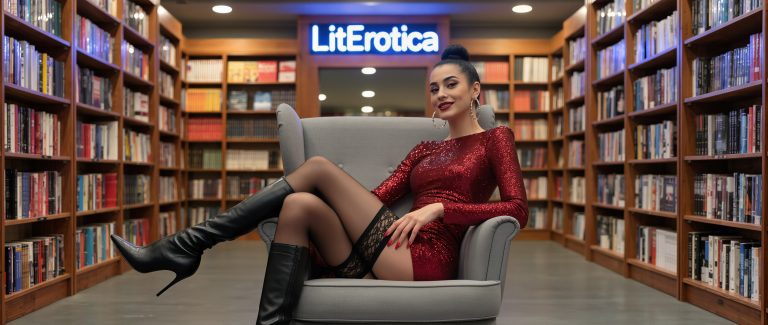 A woman in a red dress sits on a gray chair in a library, with bookshelves and a “LitErotica” sign in the background, ready for a Naomi Matthews erotic fiction author interview.
