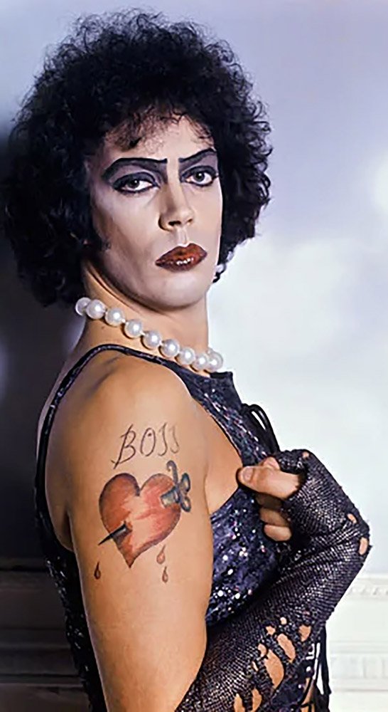 Person with dramatic makeup, curly hair, and a string of pearls poses, revealing an arm tattoo of a heart with “BOSS” and an arrow through it. Wears sequined attire and fingerless fishnet gloves.